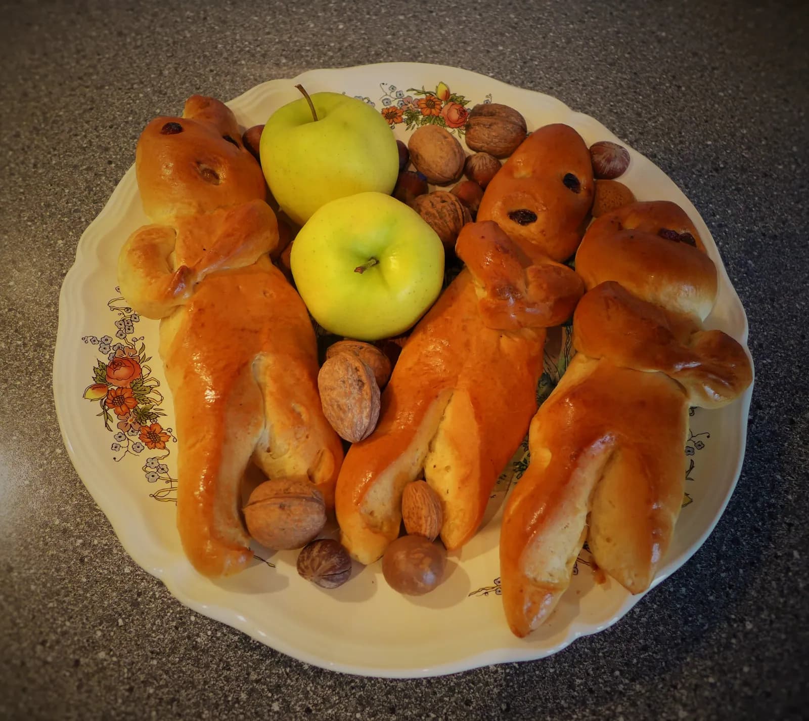 Assiette fleurie garnie de petits bonshommes en pâte briochée, de deux pommes vertes, de noix et de noisettes, posée sur un plan de travail gris
