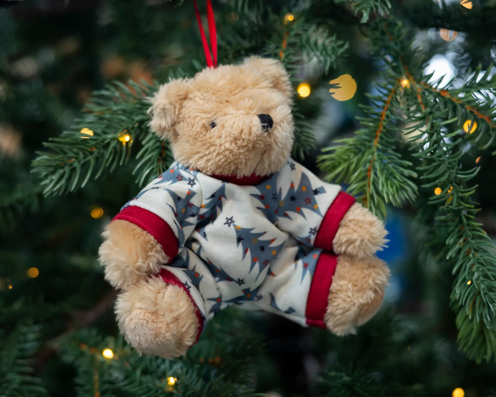 Ourson en peluche vêtu d'un pyjama blanc à motifs de sapins, suspendu à une branche de sapin de Noël illuminée