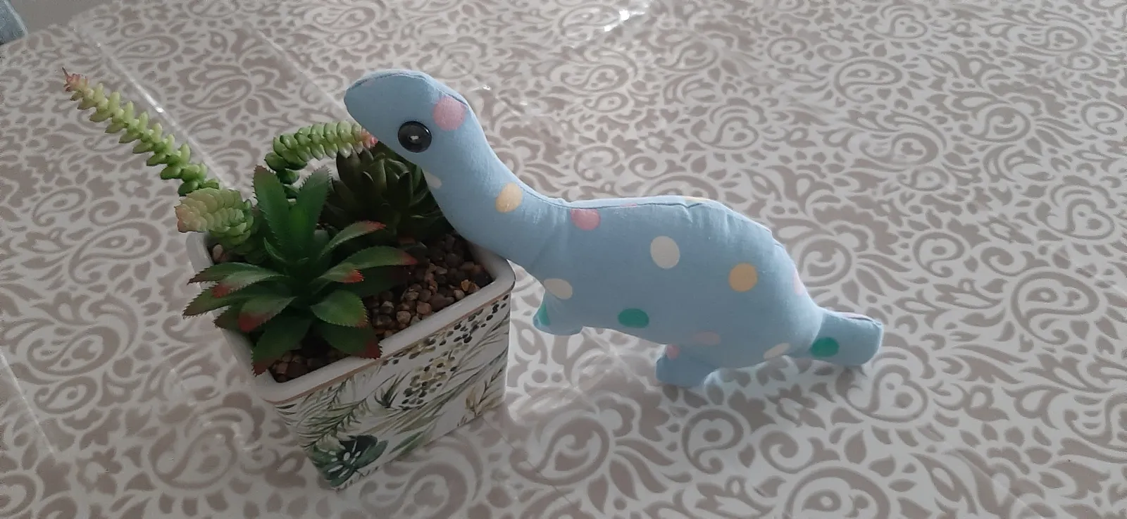 Petit dinosaure en tissu bleu clair à pois multicolores, au long cou, penché sur un pot de plantes grasses posé sur une nappe