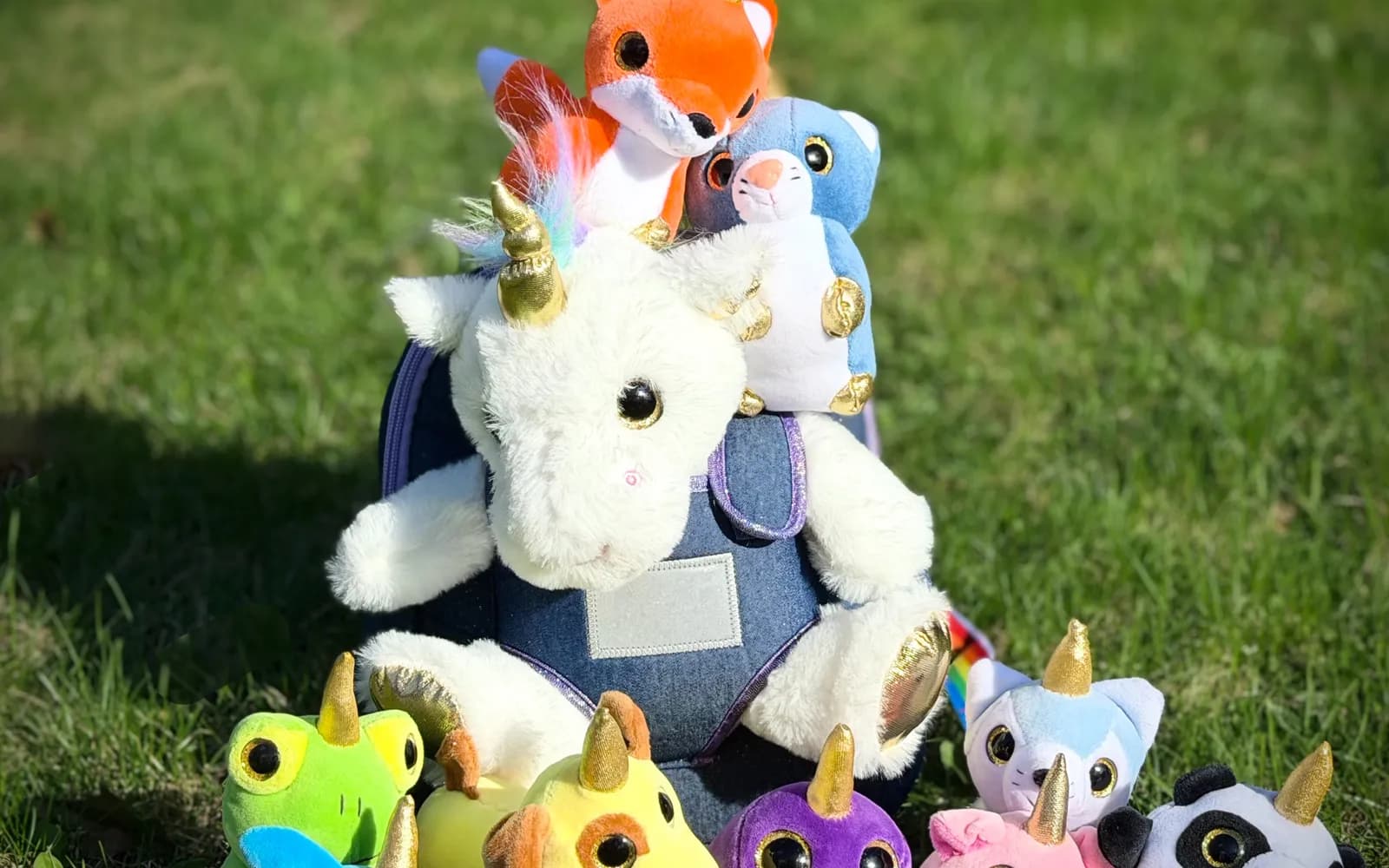 Peluche de licorne blanche à corne dorée entourée de petites peluches à corne (renard, grenouille, panda, cochon) posées sur une pelouse au soleil