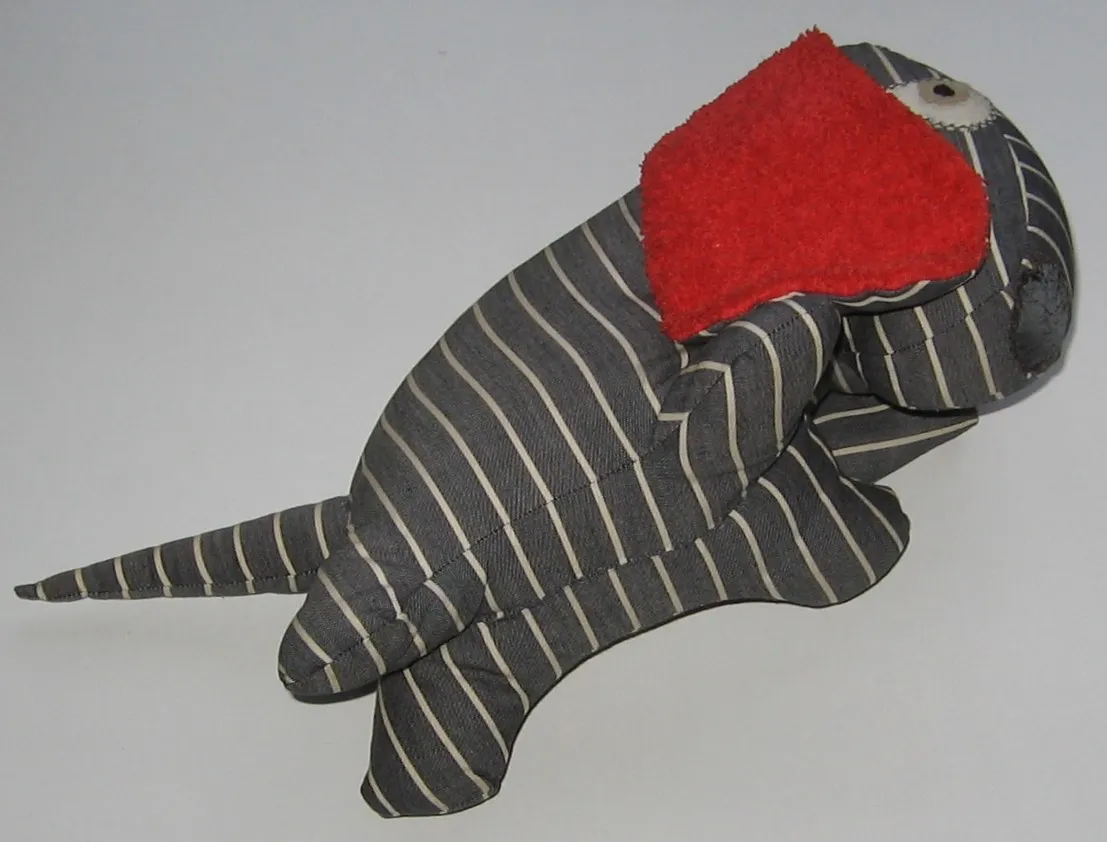 Doudou en tissu rayé gris en forme de chien, couché sur le côté, une grande oreille rouge en éponge rabattue sur la tête